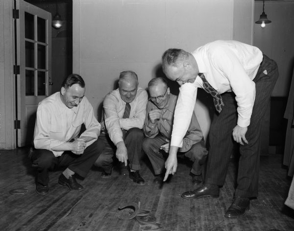 Three men are squatting on the floor, and another man is standing and pointing at rubber horseshoes. Left to right are: Arlo Anderson, Paul Steul, Al Hoessler and H.C. Timmerlake. At the annual Inter-Club Olympics, six Madison civic clubs competed in 21 events. The six clubs were the Rotary, Kiwanis, Lions, Exchange, Gyro, and Optimist Clubs.