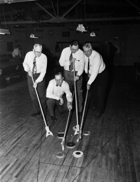 Four men holding shuffleboard push sticks over disks within a shuffleboard triangle. The shuffleboard competitors pictured are, left to right: Ray Lange, Jake Beveridge, Mo Smith and Arnie Gehner. At the annual Inter-Club Olympics, six Madison civic clubs competed in 21 events. The six clubs were the Rotary, Kiwanis, Lions, Exchange, Gyro, and Optimist Clubs.