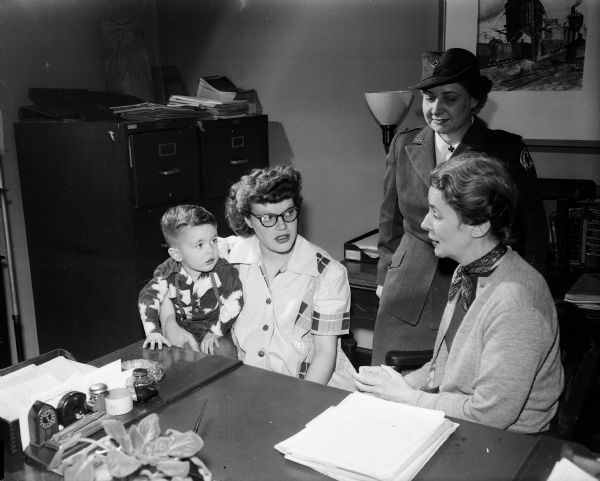 The work of the Red Cross in time of peacetime disaster is one of its least publicized services. Pictured are Mrs. Dale (Rose) Gibbs, one of the victims of a November 26, 1948 fire on Bedford Street, shown visiting the Red Cross offices with her three-year-old son, Steven, to share her experiences receiving aid from the Red Cross during that disaster with Miss Lois Pate, center, field director, and Mrs. Betty Hurst, right, home services director, both of the Dane County Red Cross at Truax Field.