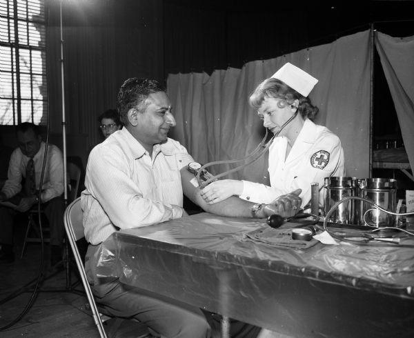 The work of the Red Cross in peacetime is less well publicized than its services during wartime. Their blood bank program is considered one of the most outstanding. Pictured is Patricia Benson, a Red Cross staff nurse, shown taking the blood pressure of one of the many donors, Dore Srinivasacher of Bangalore, India, a Ph.D student at the University of Wisconsin.
