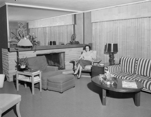 Interior view of the living room in the home of builder Tom McGovern and his family at 25 Hiawatha Circle which is decorated in shades of green and brown. A Tennessee stone fireplace fireplace occupies one wall. A mirror fills the space from the top of the fireplace to the ceiling. Mrs. Ruth McGovern is shown seated on a chair next to the fireplace