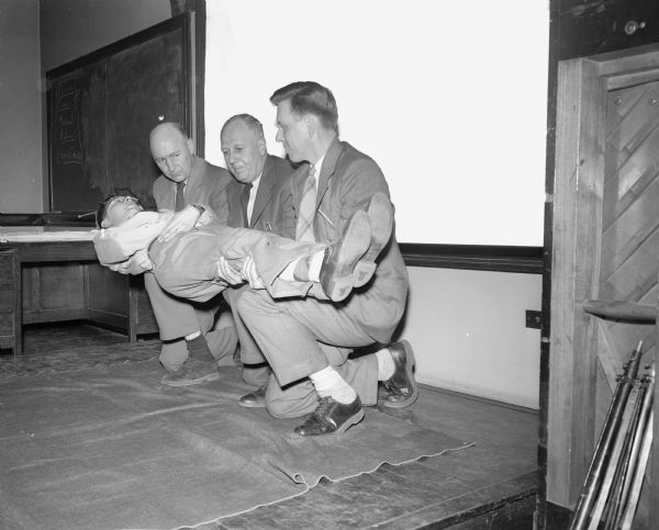 Madison teachers participating in first aid training by the Red Cross for the civil defense program. Demonstrating how to carry an injured person are Floyd Ferrill, Lawrence Johnson, Harold Foote, Ray Stesielak at East high school.