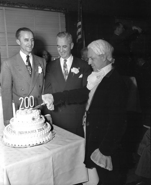 Mutual insurance men marked the 200th birthday anniversary of mutual insurance in America.  Pictured left to right are: Nels Lerdahl, program chairman, Erwin A. Gaumnitz, assistant dean of the University of Wisconsin commerce school and "Benjamin Franklin," portrayed by Walter Malec, chief accountant for Rural Mutual Insurance Company and president of the Madison Little Theater Guild, standing around the birthday cake.