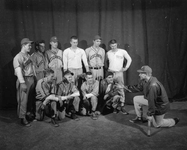 Ten members of the UW baseball team are shown listening to their coach (kneeling at right). Four players are kneeling in front and six are standing in back. Most are wearing baseball uniforms and high-top canvas shoes.