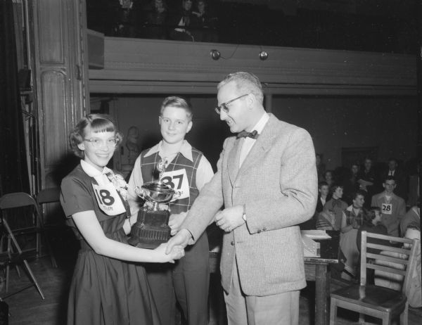 Champion Beth Knope, winner of the 1952 Badger Spelling Bee, is shown, left, clutching the <I>Wisconsin State Journal</I> trophy and shaking hands with Journal editor Roy L. Matson, right, who presented it to her.  Looking on in the middle is Warren Wickersham, Evansville champion who was runner-up in the Badger Finals.