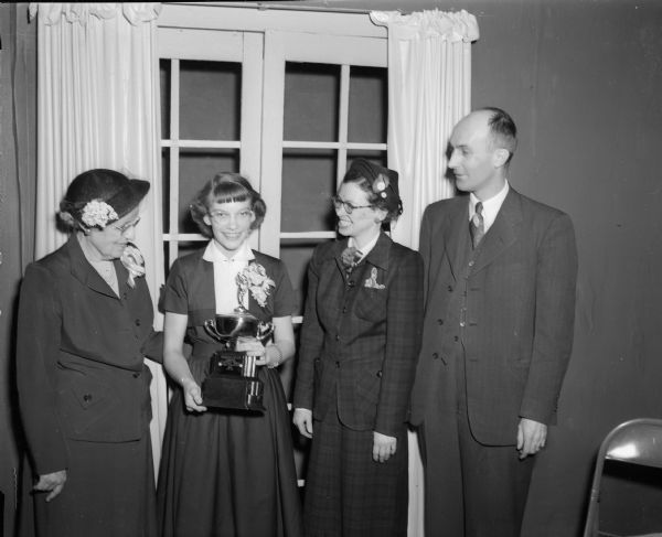 Group portrait of Beth Knope, winner of the 1952 Badger Spelling Bee, holding her trophy and standing between her teacher Floy Kendrick, on the left, and her parents Pearl and Sidney E. Knope.