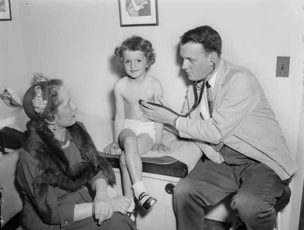 Dr. A.M. Iams (right), Madison pediatrician, uses a stethoscope to examine Julie Conlin (center), so she will be prepared to enter kindergarten. Her mother, Bernice Conlin, is sitting and watching at the left.