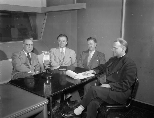 Four pastors are shown seated around a table with a radio microphone in the studio of WIBA-FM and WIBA-AM. They are planning a program that will be on the air at 10:15 each weekday night entitled "Your Pastor Speaks" that will discuss, without using a doctrinal approach, everyday living and man's brotherhood in the light of his own faith. Shown left to right, are the Rev. Roy W. Zimmer, Christ Presbyterian church; the Rev. R. G. Borwardt, Trinity Lutheran church; the Rev. Charles R. Bell, First Bapist church and the Rev. Andrew Breines, representing the Madison Catholic diocese.
