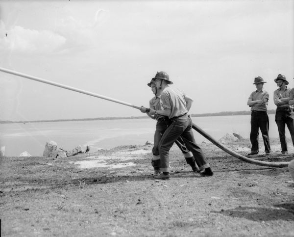 Two fire fighters are shown holding a hose with liquid shooting out of its nozzle while two other fire fighters look on. Behind them is a lake.