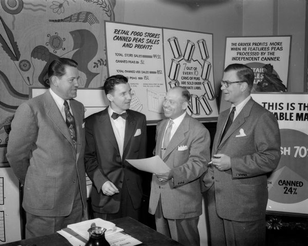 Four men in suits are posed in front of promotional posters. The newspaper caption reads: "four of the nation's leading marketing experts who have been conducting a three-day sales training institute for the canning industry at the University of Wisconsin School of Commerce are shown. They are, left to right, Watson Rodgers, president of the National Food Brokers Association,Howard Stier, director, division of statistics, National Canners Association, Roy Harb, national sales service manager, Red and White Corp., Chicago and Harold Jaeger, director of the marketing bureau of the Can Manufacturers Institute."