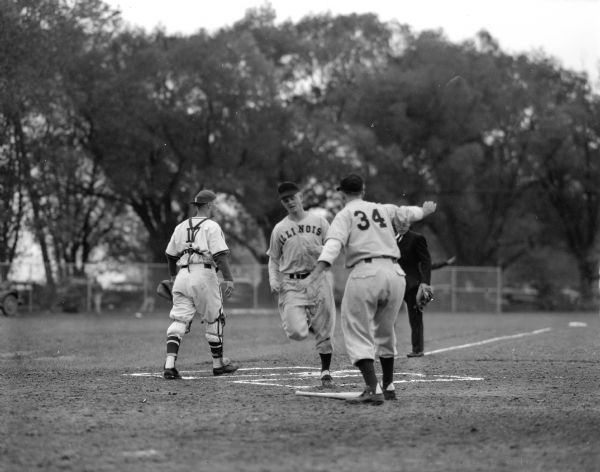Illinois player John Davis scoring a run against Wisconsin with teammate Ron Heberer greeting him. The Wisconsin catcher is Tom Cooper and the umpire is Danny Deevers. This was the first game played at the new baseball diamond on Walnut Street near University Bay.