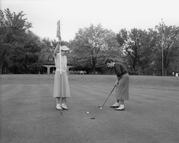 Two women golfers on the 18th green at Nakoma Country Club.  Holding the flag is Lora Krutz, women's golf chairman.  Putting is Elynore Wegner, club women's champion.