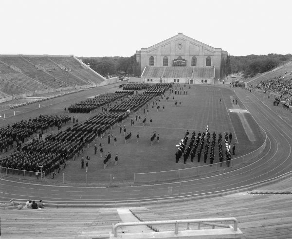 The Brigade of University of Wisconsin ROTC cadets massed in battalion fronts in Camp Randall stadium as they await the order to "pass in review" at a federal inspection. The 3,3000 cadets in the picture represent one of the largest university ROTC units in the country.  Army ROTC troops are in the left front, the midshipmen of the Naval ROTC can be identified by their white caps, and the Air ROTC cadets are at the far end of the brigade line.  Music for the review was provided by the university band, right front.