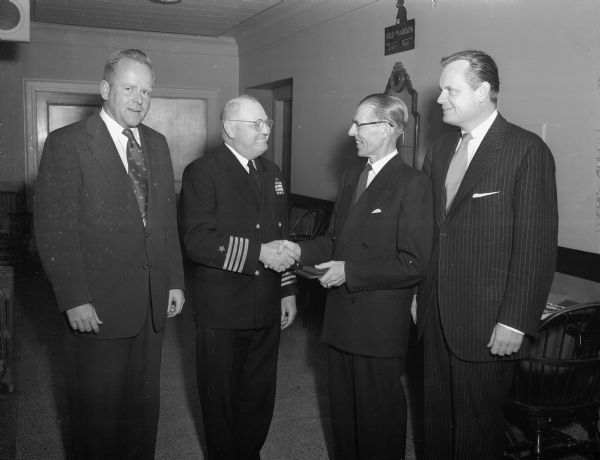 Captain Robert E. Blue, commandant of the University of Wisconsin Naval ROTC, receiving a retirement present at a banquet in his honor which was sponsored by the Madison council of the Navy League.  He is shown receiving the gift and a farewell handshake from Capt. L.K. Pollard of the Navy League. Left to right are: John G. Jamieson, Capt. Blue, Pollard, and Lawrence J. Fitzpatrick.