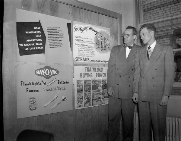 Two men standing by examples of ads, including ones for Ray-o-Vac batteries and Strauss Printing, at an Advertising Club meeting/conference.
