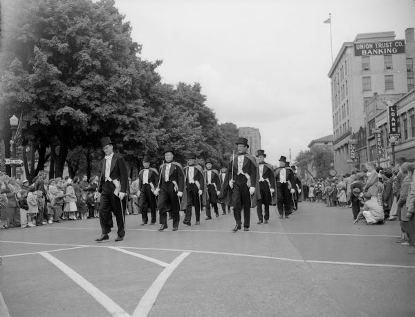 The Madison 4th degree team of the Knights of Columbus, wearing formal clothing with top hats, vests and capes and carrying walking sticks, marching in formation on West Main street.