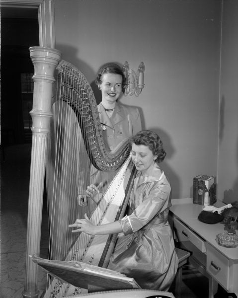 Two women at the Tri Delta "Pansy Breakfast". The purpose of the breakfast is to honor engaged and "pinned" women. Marilyn McCormick, Alpha Gamma Delta, is playing a harp, and standing by is Mary Cunnien, Sigma Kappa.