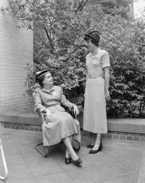 Two women visiting on the porch prior to the Tri Delta "Pansy Breakfast". Seated is Joanne Conlin, Alpha Phi, and standing is Pat Devine. The purpose of the breakfast is to honor engaged and "pinned women.