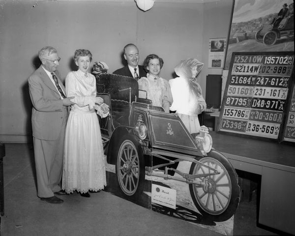 A scene at the golden jubilee open house of the Wisconsin Division of the American Automobile Association. Standing next to a 1902 Rambler automobile are staff members dressed in vintage clothes. Left to right: Ralph Hult, Madison automobile dealer; Marybelle Viney; Stuart B. Wright, general manager of the division; and Winnifred Westphal.