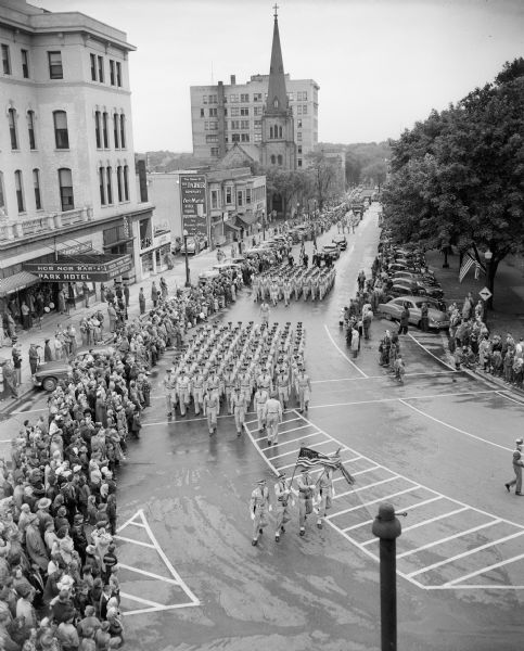 Elevated view of part of the crowd watching as military units from Truax Field march down South Carroll Street during the Memorial Day parade at Madison's Capitol Square. The Park Hotel is on the left.