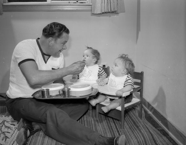 Stanley N. Davidson, 5670 Lake Mendota Drive, is shown sitting on the floor while feeding his twin sons, Dannie and Donnie. The photograph was part of a newspaper feature on fathers and their children.