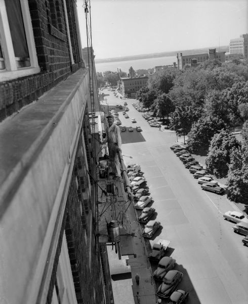 William F. Guethlein, Sauk City, resting on a scaffold on the Belmont Hotel, 31 North Pinckney Street. He has been repairing and painting the hotel windows. The Capitol Square grounds and Lake Monona are seen in the background.