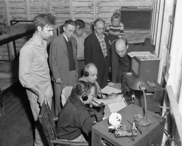 Seven members of the Four Lakes Amateur Radio Club operating a transmitter and receiving station at the club's annual field day. Standing, left to right:  Ray Bade, Haywood Cameron, Fred Foss, Frank Grams, and Jerry Dame. Seated:  William Wollin, foreground, and Dick Gelling.