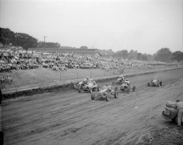 Racers coming into the first turn of the first five-mile heat at the Dane County Fair Grounds.  Shows five racers and background crowd.