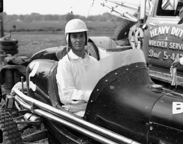Racer Bobby Grim of Indianapolis who competed in the auto races at the Dane County Fairgrounds in June, 1952. Beside him on the right is a "Heavy Duty Wrecker Service" tow truck.