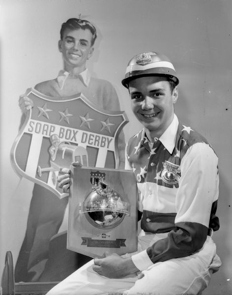 Roddy Botts, the 1951 soap box derby champion, is shown dressed in his champion's uniform and holding the trophy for the city champion in the 1952 race. In the background is a cardboard cutout of a boy holding the official derby symbol.