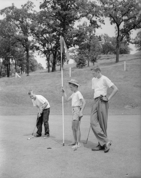 Three young golfers stand on a green during the first junior day at the Blackhawk Country Club. Putting is Dick Gregg. Ralph "Butch" Schlicht holds the flag and Fred Fitschen looks on.