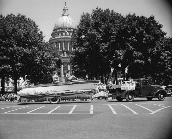 "Comely young women" ride the float of the Four Lakes Yachting Club in the 1952 Fourth of July parade on Capitol Square for the East Side Businessmen's Association. The Wisconsin State Capitol building is visible in the background.  View also includes truck pulling the float.