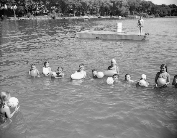 Twelve children are shown in the water at the village beach at the start of the balloon race. A boy is standing on a raft behind the group, and the shoreline is in the background.