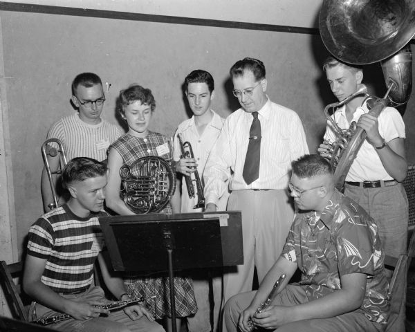 Small group portrait of six student members of the 1952 All-State high school band examining a musical score with their conductor. Shown seated at left is Gerald Buck, Lancaster; seated at right is Dick Wettstein, Madison. Standing left to right are: Gary Zwicky, Oshkosh; Darlene Knops, Waupun; Bud Bisbee, Poynette; Conductor Maurice McAdow; and David Wundrow, Middleton. The students are holding their instruments.