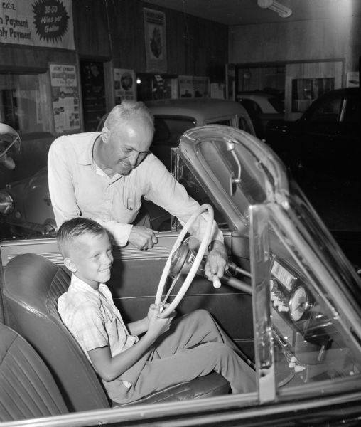 Madison Soap Box Derby entrant, 11-year-old Billy West, son of Mr. and Mrs. William West of Route 2 in Madison, is shown behind the wheel of a Willys car. O.D. Smart, president of Smart Motor Company on University Avenue and sponsor of Billy in the Derby, shows him some features of the car.