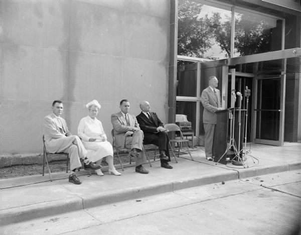 R.J. Sutherland, president of the board of directors of the Madison General Hospital Association, standing at the lectern to the left as he gives the main speech at the dedication ceremonies of the new $2,750,000 addition to the hospital. Seated left to right waiting their turn at the lectern are Mayor George Forster, retiring hospital administrator Grace Crafts, and David C. Reynolds, new hospital administrator. The new addition is in the background.