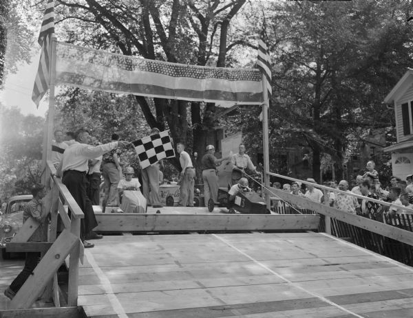 The starter, Glen (Pat) Holmes ,is holding the checkered flag at the starting ramp of the Soap Box Derby on Gorham Street as two cars prepare to take off. Holmes is the Madison recreation director.