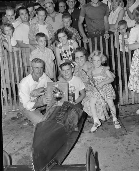 Madison Soap Box Derby winner Larry Laverty sits in his racer while holding his trophy. His family is gathered around him. The race was held on East Gorham Street. Larry's brother David, standing behind him, went on to win the 1955 Madison soap box derby.