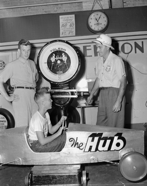 Dick Dummershausen, seated in his racer, is weighed prior to the Madison Soap Box Derby. The official weighmaster is Joe Marx (right) from Toledo Scales Company who provides the official derby scales. At left is Bob Schmitz of the Hub, Dick's sponsor.