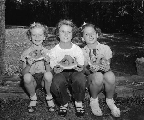 Three campers sit on a log while eating watermelon after a treasure hunt at the Brownie Hoyt Park Day Camp.  Left to right: Elizabeth O'Neill, Oregon; Bonnie Bernards, Waunakee; and Jean Topham, Oregon.