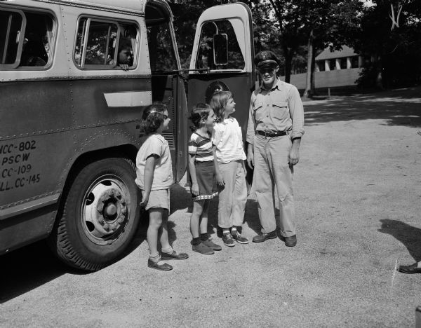 A school bus driver and three campers are standing next to the bus which transports campers to and from the Brownie Day Camp at Hoyt Park. Left to right: Susan Anderson, Sue Laugen, Cynthia Fink and driver, Bill Olsen.