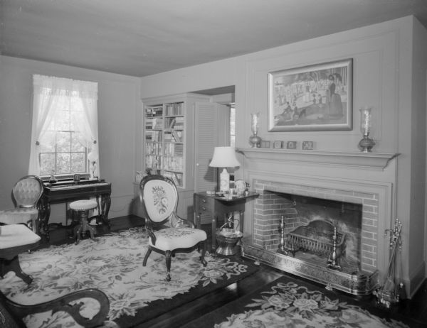 Living room with fireplace and needlepoint chair in the Elliott & Jennie Kiser house on Old Middleton Road near Heim Avenue, built in 1860 by Joseph Heim. The walls are painted Williamsburg green. At the windows are handwoven basket-weave curtains made especially for the Kisers in Virginia. The Persian copper and brass lamps on the mantel were sent from London by Kiser during the war. A reproduction of the painting by Georges Seurat "A Sunday on La Grande Jatte" is above the fireplace.