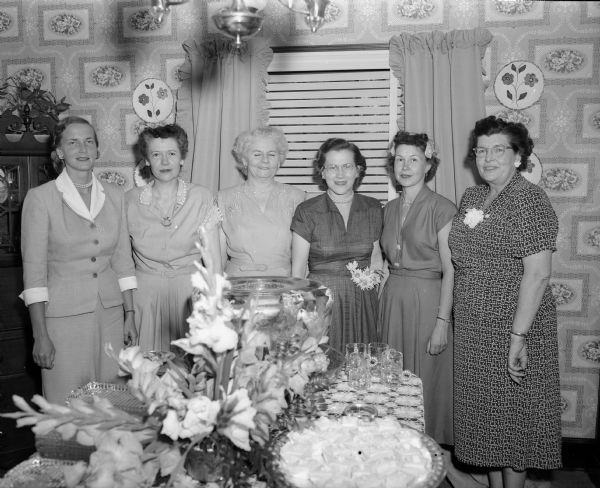 Six members of the Wisconsin Veterinary Medical Association Auxiliary are standing in front of a table prepared for a reception during a conference of the organization. Left to right: Mrs. D.K. Sorensen, general chairman of the conference; Irene Reading, state auxilary president; Mrs. F.W. Milke, Milwaukee, past president; Marcella Candlin, chairman of the tea; Mrs. A.M. McDermid, luncheon chairman; and Helen Winner, assistant publicity chairman.