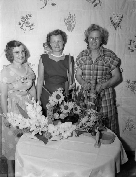 Three women are standing at a table decorated with floral arrangements at the Mendota Gardeners garden show. Left to right: Ellen Antoine; Marie Connor, president of the garden club; and Mrs. Troy Gordon.