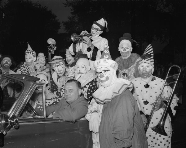 Clowns surround Mayor George Forster, sitting the the driver's seat of a car, during the Knights of Columbus Shrine Softball Circus event.