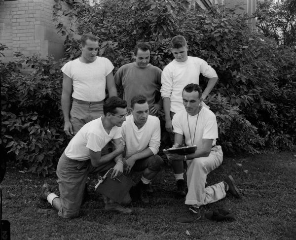 Group portrait of Wisconsin High School football coaching staff looking at a clip board. Shown knelling in the front row are, Pete Kintis, Ronald Bailey, and Coach Hal Metzen. In the back row are Don Damon, Dick Trotta, and Robert Blue.