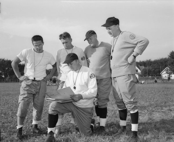 Outdoor group portrait of five football coaches at East High School. Standing left to right: assistant coaches Jerry Butterfield, Milt Diehl, Ferris "Red" McKinley, and Larry Johnson. Kneeling is Coach Herbert "Butch" Mueller.