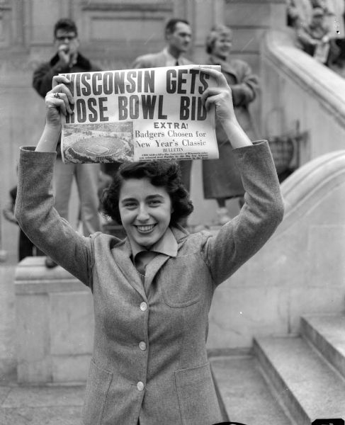 A female student holds up "Capitol Times" newspaper of November 24, 1952 announcing University of Wisconsin-Madison's bid for Rose Bowl participation. Other people are standing in the background in front of what appears to be possibly Memorial Union or the Capitol.