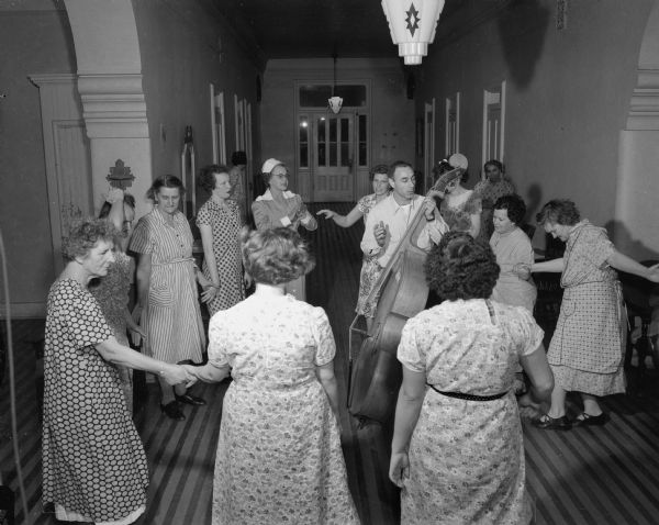A circle of female patients and a nurse hold hands during a dance at Mendota State Hospital. John Birchall, a hospital recreation staff person, plays a double bass at center.