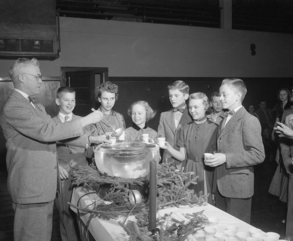 Nakoma Elementary School children standing around a punch bowl at a holiday party sponsored by the Nakoma Welfare League. League member Wilmer Ragatz, left, is pouring the punch. Students left to right: Gary Reiman, Margie Pallett, Terry Thomas, Bob Wright, Virginia Ward, and Stewart Honeck.